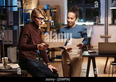 Contenu jeunes collègues multiethniques analysant des graphiques tout en travaillant avec des documents au bureau la nuit Banque D'Images