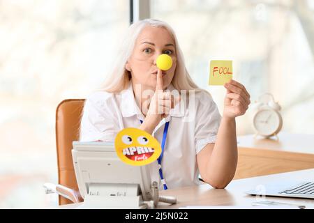 Drôle de femme mûre avec le nez clown et le papier collant montrant le geste de silence dans le bureau. Fête de la journée d'avril Banque D'Images