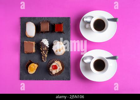Plusieurs gâteaux sur une plaque de pierre rustique à côté de quelques tasses de café, fond rose. Banque D'Images