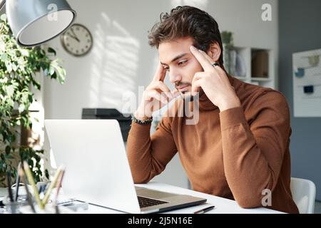 Un jeune homme sérieux du Moyen-Orient en turtleneck brun s'est concentré sur les pensées regardant l'écran d'ordinateur portable et les temples massants Banque D'Images