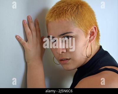 Jeune femme alternative attrayante d'hipster mexicain Latina avec des cheveux courts teints à l'orange et regarde curieusement sur les côtés le spectateur devant un mur gris. Banque D'Images