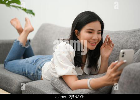 Charmante jeune femme asiatique se détend à la maison, se reposant sur un canapé confortable et profitez des appels vidéo avec ses amis via un smartphone. Banque D'Images