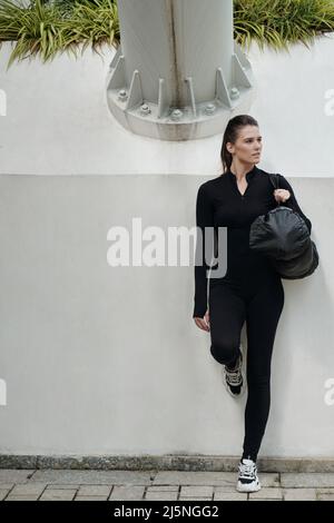 Jeune femme en dancewear tenant un sac de gym et s'appuyant sur le mur dans le parc Banque D'Images