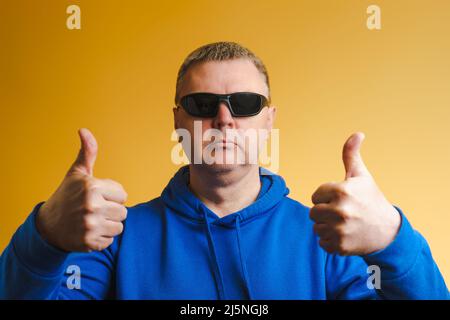 Portrait d'un homme dans un sweat à capuche bleu et des lunettes de soleil. Un homme montre un geste de pouce vers le haut sur les deux mains. Banque D'Images