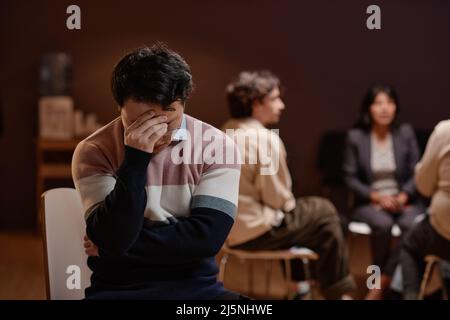 Portrait horizontal moyen d'un jeune homme asiatique adulte souffrant de dépression prenant part à la thérapie de groupe assis sur la tête d'abaissement de chaise Banque D'Images