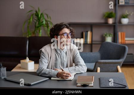 Portrait horizontal moyen d'un jeune adulte élégant portant une tenue décontractée élégante avec des lunettes assis sur le bureau et donnant sur le côté Banque D'Images