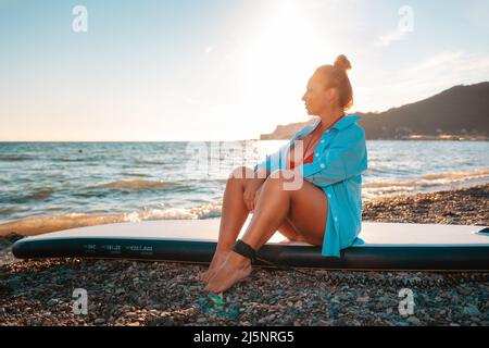 Une femme caucasienne en chemise pose assise sur un plateau de sup. Coucher de soleil et océan en arrière-plan. Le concept de la récréation sportive au bord de la mer. Banque D'Images