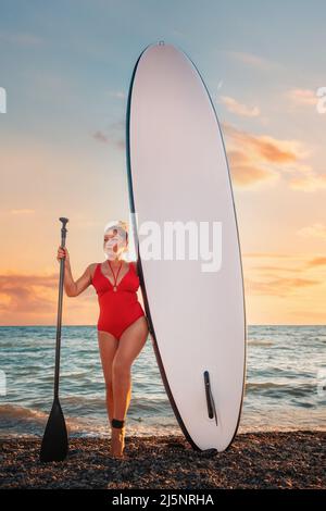 Femme caucasienne adulte Plump dans un maillot de bain rouge tenant un panneau supérieur et paddle. Coucher de soleil sur l'arrière-plan. Orientation verticale. Le concept d'actif Banque D'Images