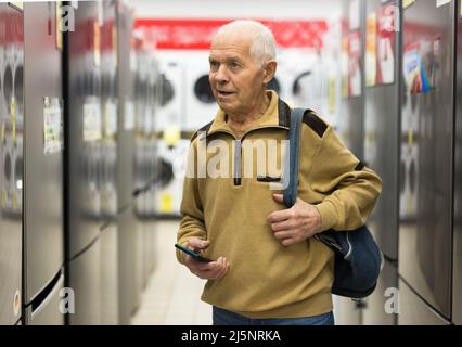 homme âgé à la recherche d'un réfrigérateur au comptoir dans la salle d'exposition du service d'hypermarché des appareils électriques Banque D'Images