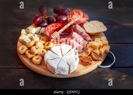 Planche à découper ronde avec assortiment de tranches de fromage, rosette à saucisse, jambon, craquelins, raisins et figues au miel. Assiette de charcuterie et de fromage sur le plat Banque D'Images