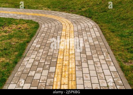 Chemin traversant la pelouse dans le parc. Revêtement tactile jaune vif pour les personnes malvoyantes sur le trottoir. Banque D'Images