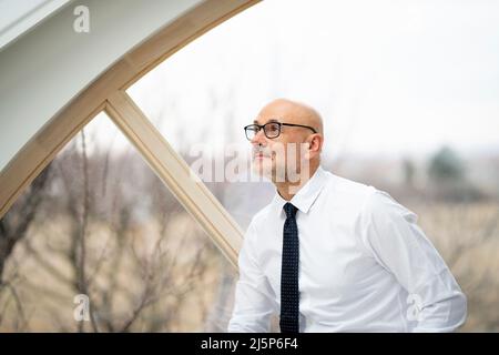 Portrait d'un homme d'âge moyen souriant portant une chemise et des lunettes tout en étant assis à la fenêtre et en regardant loin. Banque D'Images