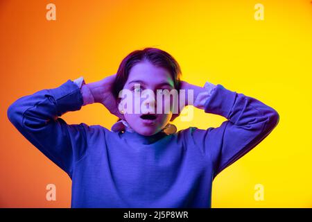 Portrait de garçon, enfant mettant les mains derrière la tête avec l'expression choquée isolé sur fond jaune studio au néon Banque D'Images