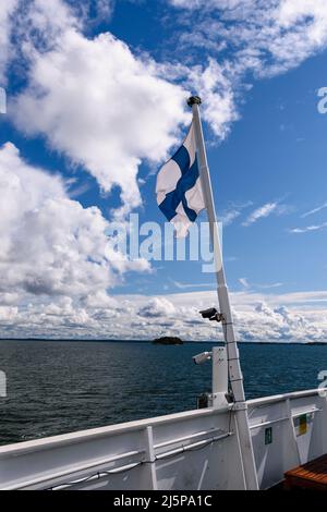 Un drapeau finlandais agitant contre le ciel bleu avec des nuages et une île en arrière-plan sur un navire dans l'archipel de Turku, en Finlande. Banque D'Images