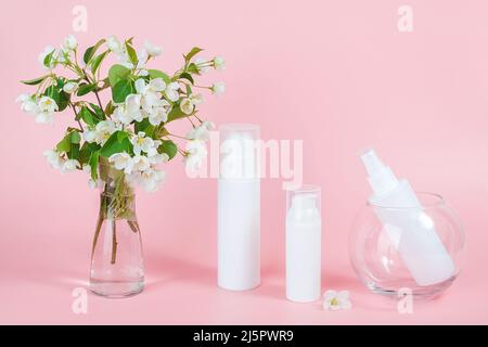 Ensemble de cosmétiques pour le soin de la peau du visage, du corps. Flacons et tube blancs pour cosmétiques sur podium en verre et branche en fleur dans vase, fond rose. Natura Banque D'Images