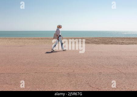 Une femme promeneuse solitaire sur une promenade au bord d'une plage de galets et d'une mer bleue calme Banque D'Images