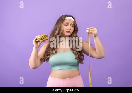 Triste malheureux affamé jeune femme européenne plus taille dans les vêtements de sport avec Burger à la main regarde le mètre ruban Banque D'Images