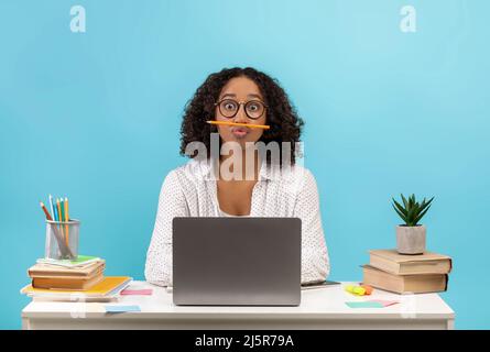 Une jeune étudiante noire stupide assise au bureau avec des livres et un ordinateur portable, jouant au jeu avec un crayon sur fond bleu de studio Banque D'Images