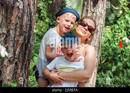 Mère et petits garçons se penchent sur des troncs de pin dans le parc de l'hôtel. Une femme en lunettes de soleil tient le tout-petit et l'enfant d'âge préscolaire dans des chapeaux le jour ensoleillé de gros plan Banque D'Images