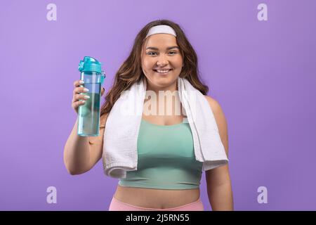 Bonne jeune femme européenne de taille plus dans les vêtements de sport avec une serviette montrant une bouteille d'eau Banque D'Images