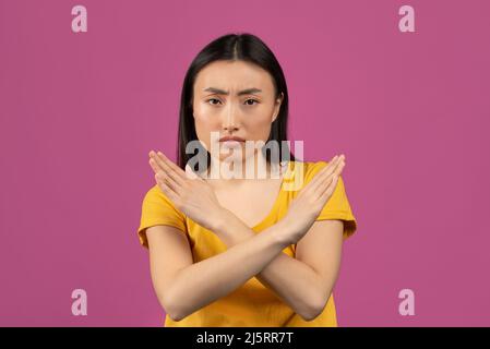 Portrait d'une femme asiatique sérieuse montrant un geste d'arrêt avec des mains croisées, posant sur fond de studio violet Banque D'Images