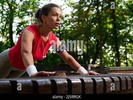 Gros plan d'une athlète hispanique féminine déterminée, une femme musclée dans des vêtements de sport brillants effectuant des push-up à partir d'un banc en bois dans le parc dur de la ville Banque D'Images