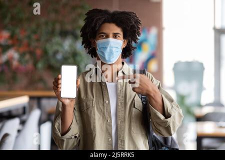Joyeux millennium afro-américain mauricien étudiant dans un masque de protection avec sac à dos pointant le doigt vers le smartphone Banque D'Images