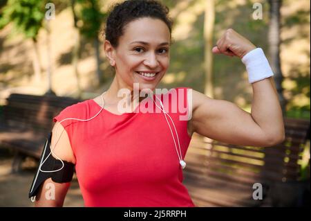 Allez au-delà de vos limites. Active multi ethnique moyenne d'âge athlète musclé sportswoman souriant beau sourire et montrant son biceps tonique à la came Banque D'Images