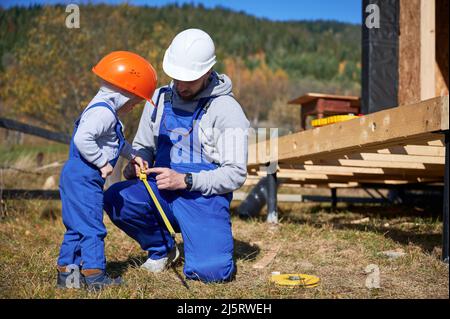 Père avec enfant fils bâtiment en bois maison de cadre. Homme de construction et enfant jouant avec mètre ruban sur le chantier, portant un casque et une combinaison bleue le jour ensoleillé. Menuiserie et concept de famille. Banque D'Images