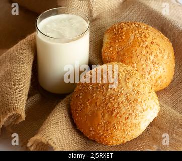 Encore la vie avec un verre de lait et deux petits pains. Un verre de lait maison et deux délicieux petits pains au sésame fraîchement cuits sont placés sur un tissu épais. Banque D'Images