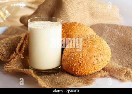 Encore la vie avec un verre de lait et deux petits pains. Un verre de lait maison et deux délicieux petits pains au sésame fraîchement cuits sont placés sur un tissu épais. Banque D'Images