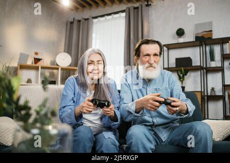 Bonne bonne vieille femme et son mari barbu tenant la console de jeux tout en étant assis ensemble sur le canapé. Loisir avec plaisir d'aimer les vieux, la retraite. Banque D'Images