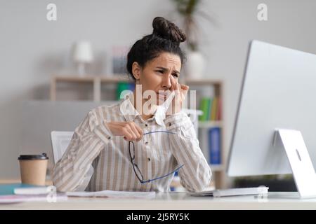 Une femme ravie, malheureuse et fatiguée, se frottant les yeux et pleurant, assise sur le lieu de travail à l'intérieur du bureau avec un ordinateur Banque D'Images