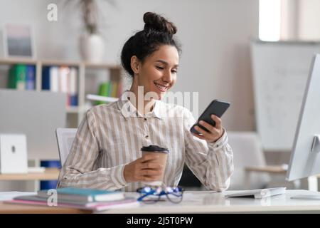 Une femme heureuse utilise son smartphone et boit du café au bureau, se fait une pause au travail, s'assoit au bureau et dispose d'espace libre Banque D'Images