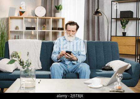 Les gens, la retraite, la maison et l'utilisation de gadgets. Homme barbu senior, assis sur le canapé dans un salon confortable à l'intérieur et utilisant un smartphone pour écrire des messages ou des serfing réseaux sociaux. Banque D'Images