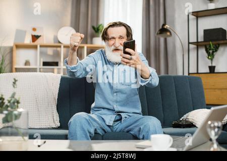 Les gens à la maison et en utilisant des gadgets. Homme plein de style et plein de confiance avec une barbe grise souriant assis confortablement sur le canapé dans un salon confortable à l'intérieur et utilisant un smartphone pour écrire des messages ou des serfing réseaux sociaux Banque D'Images