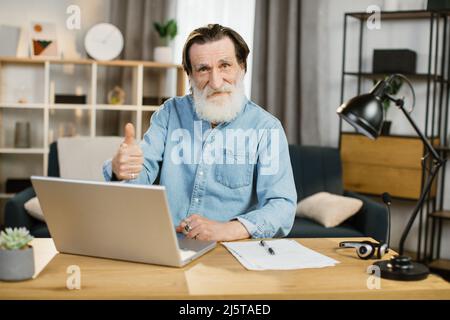 Beau développeur de logiciels caucasiens senior dans la dactylographie de port occasionnel sur ordinateur portable tout en étant assis sur le lieu de travail et en montrant le pouce vers le haut. Employé de bureau très occupé homme mûr utilisant des gadgets modernes pour le travail Banque D'Images