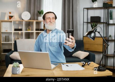 Homme d'affaires mûr souriant utilisant un smartphone moderne pour réaliser des photos de selfie. Homme plein d'assurance avec barbe grise assis à table avec ordinateur portable et papiers. Technologie, fun, travail à distance. Banque D'Images