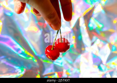 Flou artistique main tenant deux cerises rouges mouillées avec queue et feuilles sur fond hologramme moderne. Fruits et baies. Récolte et conc. De récolte Banque D'Images