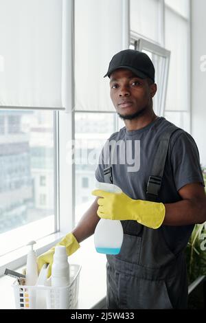 Jeune Africain américain contemporain avec détergent en bouteille plastique debout près du seuil avec récipient tout en lavant les fenêtres Banque D'Images