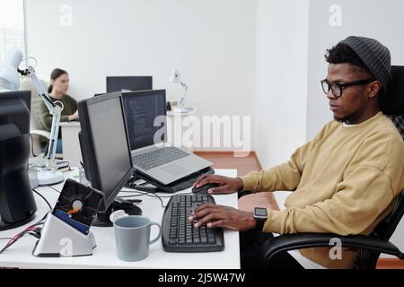 Jeune développeur américain d'informatique en mode décontracté, décodage des données tout en étant assis devant un écran d'ordinateur dans un bureau moderne Banque D'Images