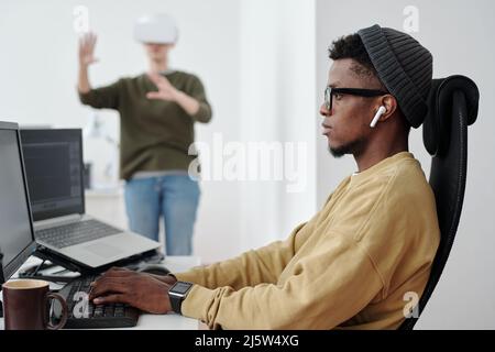Vue latérale d'un jeune homme d'affaires sérieux dans des écouteurs, des lunettes et des données de décodage de vêtements décontractés tout en regardant l'écran d'ordinateur Banque D'Images
