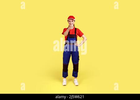 Portrait complet de femme souriante satisfaite travailleur debout et pointant le doigt vers l'appareil photo, vous choisir, porter une combinaison et une casquette rouge. Studio d'intérieur isolé sur fond jaune. Banque D'Images