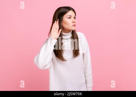 Curieuse femme avec des cheveux foncés tenant la main près de l'oreille et écoutant quelqu'un conversation, secrets, portant blanc style décontracté pull. Studio d'intérieur isolé sur fond rose. Banque D'Images