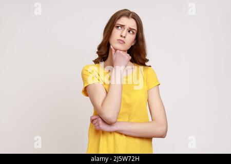 Portrait d'une jeune fille attrayante avec des cheveux ondulés bruns dans un T-shirt jaune pensant à des choses importantes, tenant le menton, regardant loin. Prise de vue en studio isolée sur fond gris. Banque D'Images