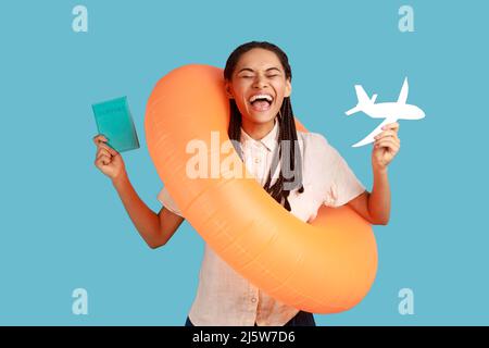 Une femme stupéfaite tient son passeport avec des cartes d'embarquement et un avion en papier, a un voyage d'été, hurler joyeusement, se réjouir de son voyage, portant une chemise blanche. Studio d'intérieur isolé sur fond bleu Banque D'Images