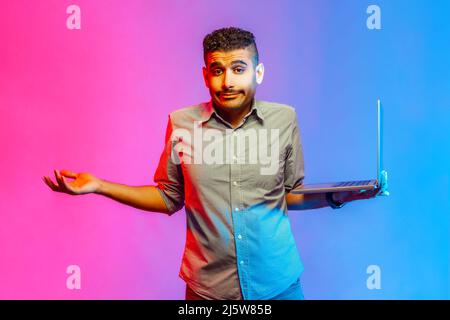 Portrait d'un beau homme perplexe dans une chemise tenant un ordinateur portable et haussant les épaules, ne sais pas quoi faire avec un nouveau projet. Studio d'intérieur isolé sur fond de néon coloré. Banque D'Images