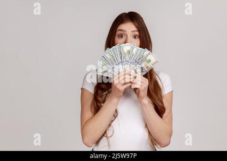 Surprise heureuse femme avec des cheveux bruns cache face derrière un fan de billets de dollars, retrait d'argent sans intérêt, portant un T-shirt blanc. Prise de vue en studio isolée sur fond gris. Banque D'Images