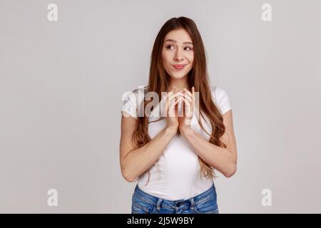 Intrigue dévot femme attrayante clasping les mains et de penser à un plan délicat, ayant une idée astucieux de se lancer, portant un T-shirt blanc. Prise de vue en studio isolée sur fond gris. Banque D'Images