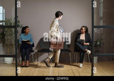 Jeune homme avec une valise assise sur une chaise entre deux femmes dans le corrifor de bureau Banque D'Images